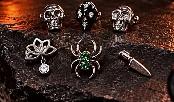 Joyería para perforación sobre piedra oscura: tres calaveras, una flor de loto con gema colgante, una araña con gemas verdes y una daga