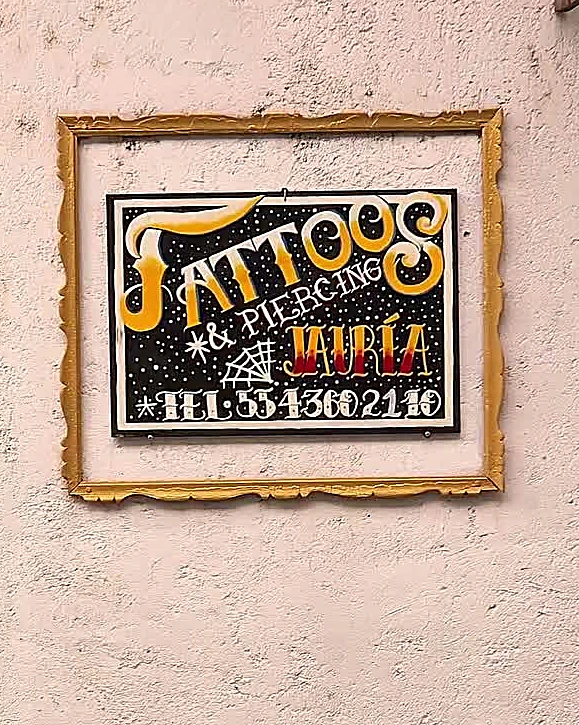 Letrero pintado a mano Tattoos y Piercing Jauría con marco dorado, telaraña y el teléfono 55 4360 2110, sobre el muro de cal de la entrada