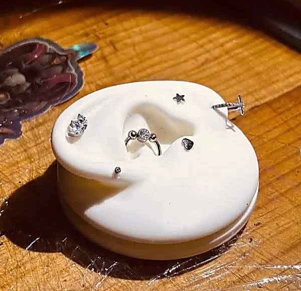 Oreja de silicón de muestra con seis piezas de joyería puestas: gema doble, estrella, cruz con brillantes, aro con gema, corazón y balín pequeño
