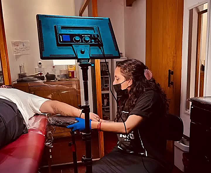 Tatuadora con cubrebocas y guantes azules tatuando un brazo junto al sillón rojo y la lámpara del estudio
